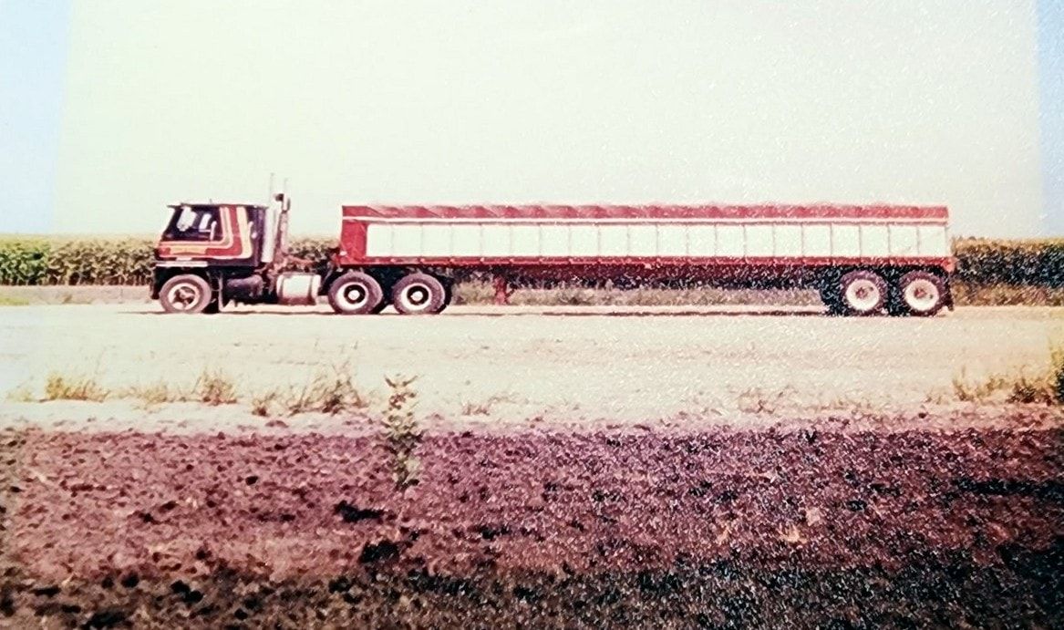 A red-striped International cabover pulling a long red grain trailer past a cornfield.