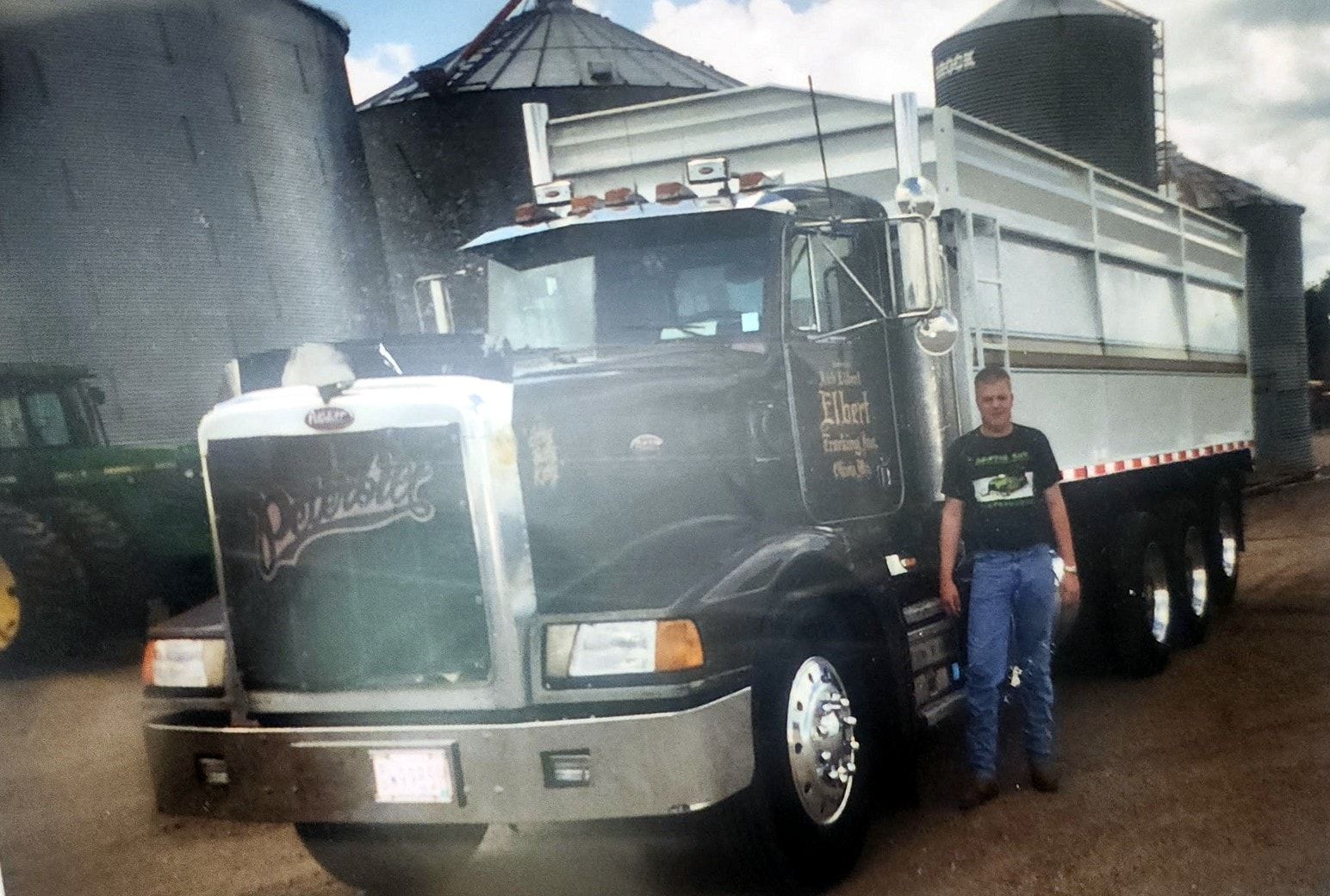 A young man standing beside a dark Peterbilt grain truck in front of grain bins.