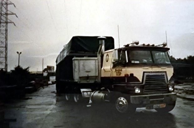 A cream and brown International cabover with a tarped load, parked on a wet lumber yard.
