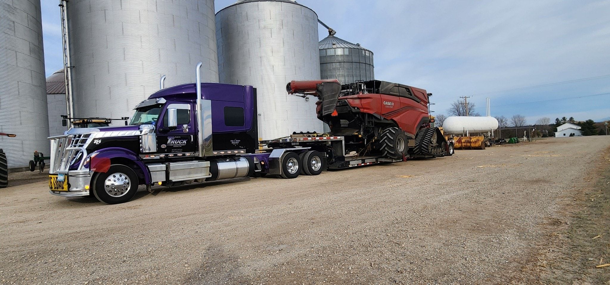 A purple and black International LoneStar hauling a red combine on a lowboy beside grain bins.