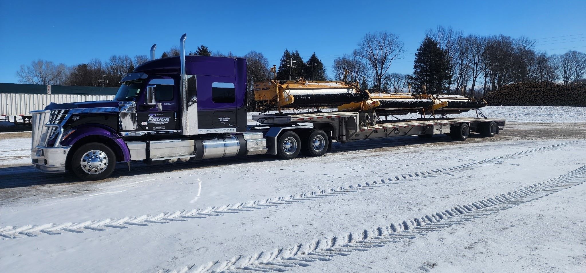 The purple and black International LoneStar on a step-deck with yellow harvester heads, in the snow.