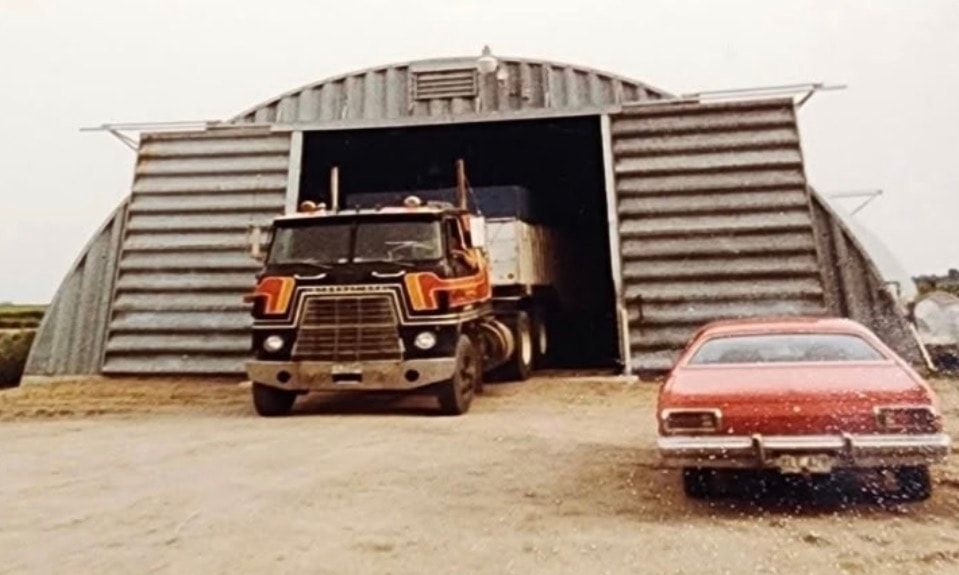 A black cabover tractor-trailer pulling out of a Quonset hut, with a red car parked in front.